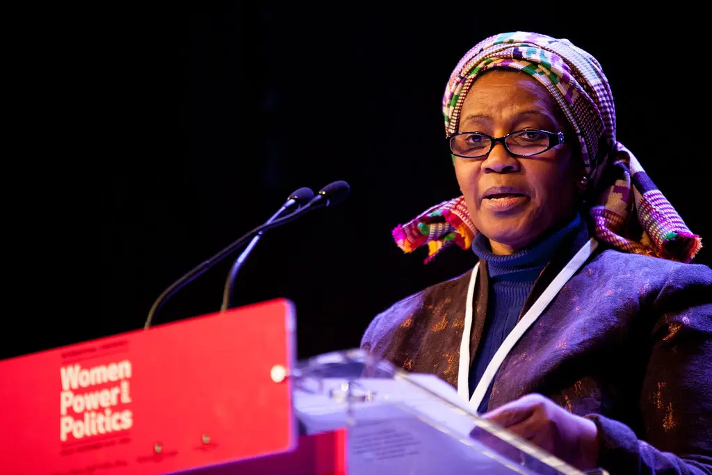 A woman wearing glasses and a colorful headscarf speaks at a podium with microphones. The podium displays a red sign reading Women Power Politics in white text.