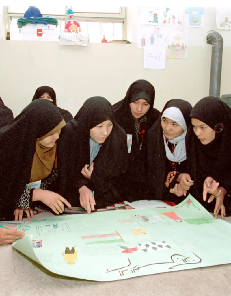 A group of girls in black hijabs sit on the floor, closely gathered around a large poster with colorful drawings and text, discussing and pointing at the project together. Classroom artwork is displayed on the wall behind them.
