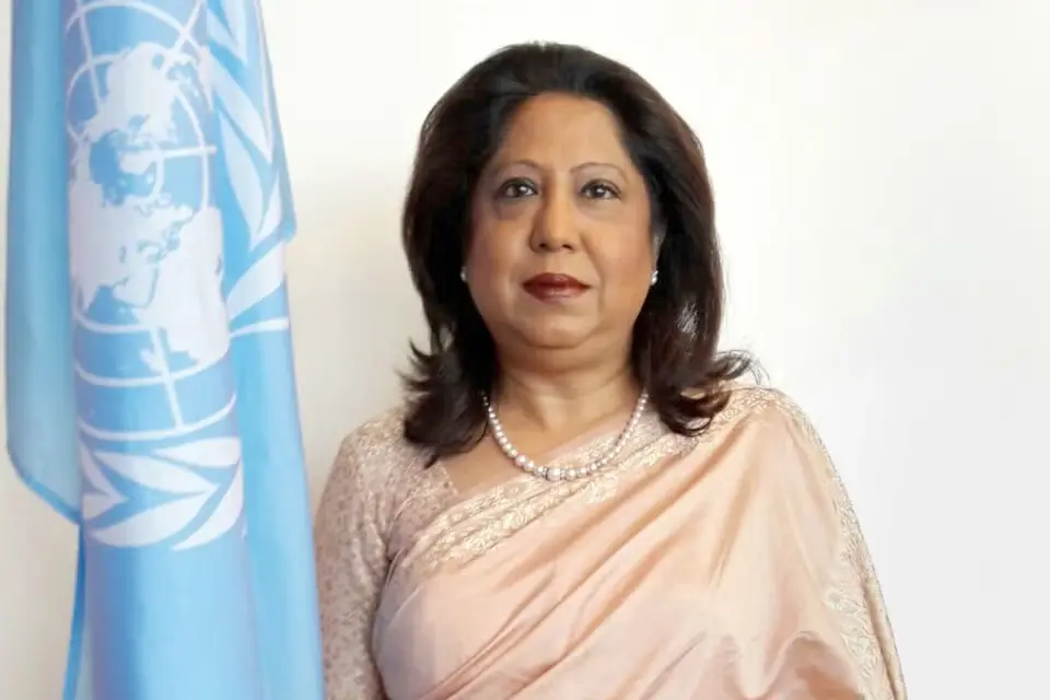 A woman in a light pink sari and pearl necklace stands next to a United Nations flag, posing against a plain white background.