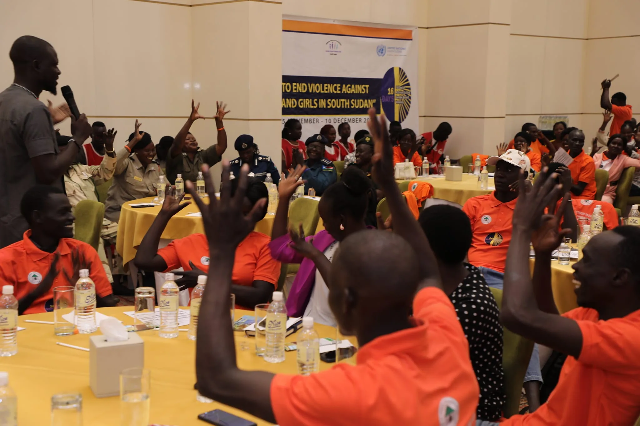 A group of people, many wearing orange shirts, sit around tables raising their hands at an indoor event. A speaker stands in front, and a banner about ending violence against women and girls in South Sudan is visible in the background.
