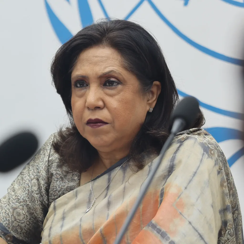 A woman with dark hair wearing a light-colored saree sits in front of a microphone, looking serious. A blue and white abstract design is visible in the background.