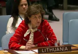 A woman in a red sweater and scarf speaks at a conference table with a nameplate reading Lithuania. She is seated with a microphone and a glass of water in front of her, with other people in the background.