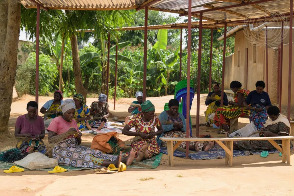 A group of women sit outdoors under a simple shelter, weaving and working with their hands. They are surrounded by trees and greenery, and appear focused on their craft, with materials and baskets around them.
