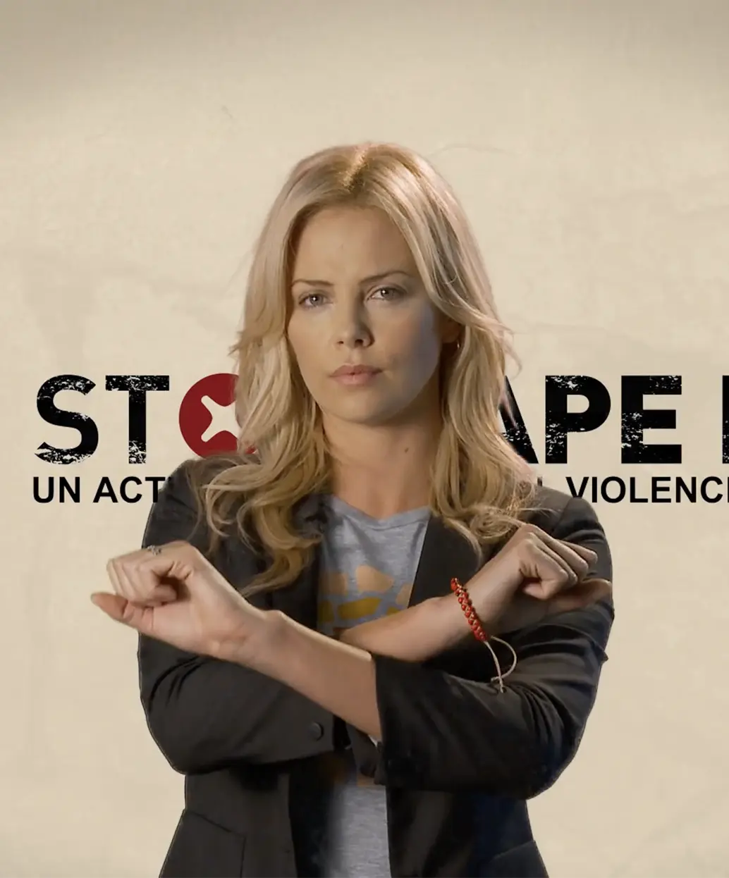 A woman with blonde hair stands in front of a sign reading STOP RAPE and holds her arms crossed in an X gesture, symbolizing protest or refusal.